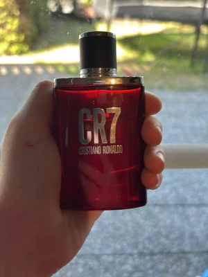 CR7 parfym från Cristiano Ronaldo - Snygg parfymflaska i röd glasdesign med silverdetaljer och svart lock. Märket CR7 är tydligt på framsidan och flaskan har en modern, stilren form. Perfekt för dig som vill ha en unik och fräsch doft i samlingen. Ca 22ml av 30ml kvar