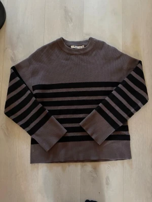 Grå och svart randig stickad tröja - Säljer en grå stickad tröja med svarta horisontella ränder över bröstet och ärmarna. Tröjan har rund halsringning och raka ärmar. 