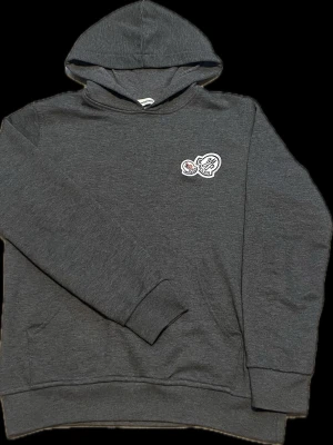 Mörkgrå hoodie med tryck - Snygg mörkgrå hoodie med huva och ett litet vitt och rött tryck på bröstet. Hoodien har ribbade muddar vid ärmslut och nederkant samt en klassisk passform. Perfekt för en avslappnad och trendig stil. OBS!Storlek M men passar S också!