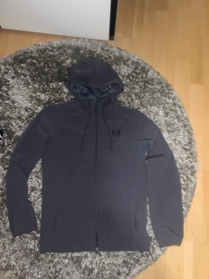 Svart vindjacka från Under Armour - Snygg svart vindjacka från Under Armour med huva och dragsko. Jackan har hel dragkedja framtill, två sidofickor och diskret logga på bröstet. Tillverkad i lätt och vindtätt material som passar perfekt för blåsiga dagar.