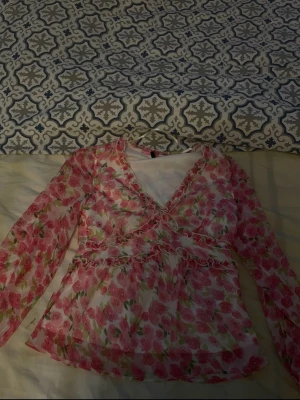 Blommig rosa meshblus med volanger - Söt blus i mesh med rosa och grönt blommönster. V-ringad framtill med volangdetaljer och markerad midja. Långärmad och lätt transparent, perfekt för en trendig och romantisk look. Materialet är tunt och luftigt, vilket ger en skön känsla.