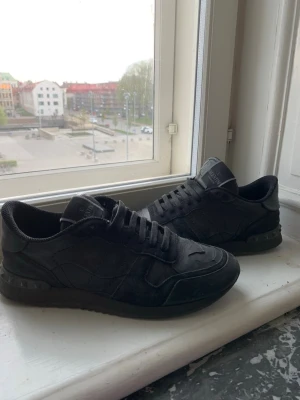 Valentino rockrunner - Snygga helsvarta sneakers från Valentino med klassisk låg siluett. Skorna har en stilren design med svarta skosnören, diskreta detaljer och ovandel i läder och textil. Perfekta för dig som gillar en clean och exklusiv look.