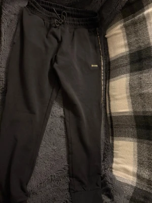 Svarta mjukisbyxor från BOSS - Snygga svarta mjukisbyxor från BOSS med dragsko i midjan och ribbade muddar vid bensluten. Byxorna har en diskret logga på vänster ben och är tillverkade i mjukt material som känns skönt mot huden. Perfekta för chill dagar eller när du vill ha en clean look.
