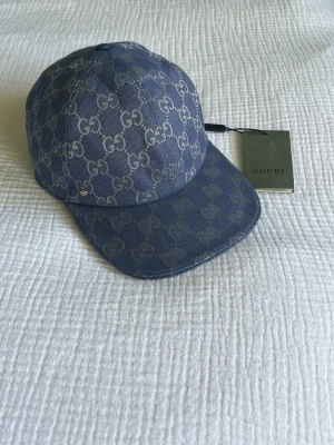 Blå keps från Gucci med GG-mönster - Snygg blå keps från Gucci med klassiskt GG-monogram över hela kepsen. Kepsen har böjd skärm och är tillverkad i canvasmaterial. Perfekt accessoar för att lyfta din streetwear-look.