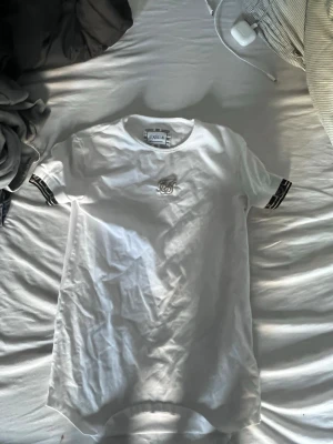 Vit t-shirt från SikSilk med logga - Vit t-shirt från SikSilk med broderad logga på bröstet och svarta detaljer på ärmsluten. Klassisk rund halsringning och normal passform. Perfekt för en stilren och sportig look.