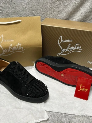 Christian Louboutin- Strl 42  - Svarta sneakers från Christian Louboutin med klassisk röd sula och svarta nitar på tån. Skorna har snörning, är låga och tillverkade i mocka och läder. Perfekta för dig som vill sticka ut med en edgy och lyxig stil. Kom privat för mer info, de är helt nya och oanvända och vi kan diskutera priset privat 