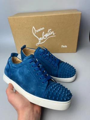 Blå Christian Louboutin sneakers mocka - Snygga blå sneakers från Christian Louboutin i mjuk mocka med matchande blå snörning och coola nitar på tån. Skorna har vit platt sula och den klassiska röda undersidan med Louboutin-logga. Perfekt för dig som gillar att sticka ut med exklusiva detaljer. Endast skor tillkommer 