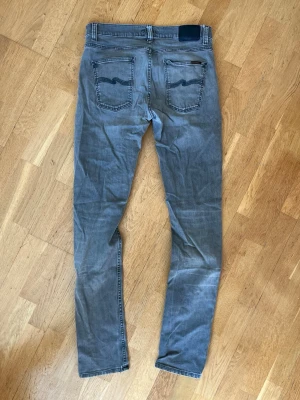 Grå slim jeans från Nudie Jeans 32/36 - Säljer ett par grå Lean Dean jeans från Nudie Jeans med klassisk femficksdesign och snygga detaljer på bakfickorna. Jeansen har en modern passform och är tillverkade i bomull med lite stretch för extra komfort. Perfekt för dig som gillar stilrena och coola jeans. Storlek 32/36