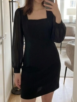 Svart klänning med transparenta ärmar - Stilren svart klänning från Lindex med fyrkantig halsringning och långa transparenta ärmar i chiffong. Klänningen har en rak passform och är tillverkad i ett mjukt material som ger en elegant look. Perfekt för dig som gillar klassiska och trendiga plagg.