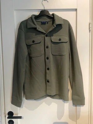 Olivgrön overshirt med fickor - Stilren olivgrön overshirt med klassisk krage och stora bröstfickor med lock. Knäppning framtill med svarta knappar och långa ärmar. Tillverkad i ett mjukt, strukturerat tyg som ger en avslappnad look. Perfekt lager-på-lager-plagg för en trendig stil.