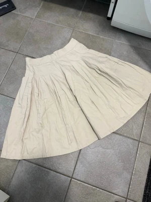 Vinted kjol med dragkedja - Snygg beige kjol med vid passform och veckade detaljer. Kjolen har en markerad midja och stängs med dragkedja bak. Perfekt för dig som gillar en klassisk och stilren look. Ej använd av mig, för liten. Står storlek 44 i den men är mer en small/medium.  Midjemått 40 cm rakt över.