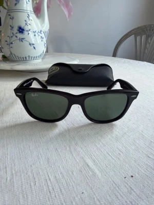 Svarta Ray-Ban Wayfarer solglasögon - Klassiska svarta Ray-Ban Wayfarer solglasögon med gröntonade glas och ikonisk bågform. Snygg och tidlös design med Ray-Ban-logga på skalmen. Kommer med originalfodral. Perfekta för dig som vill ha en cool och stilren look.