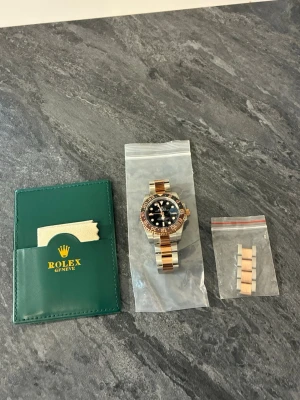 herr klocka - Säljer en herrklocka med tvåtonad länk i stål och roséguld. Klockan har svart urtavla, keramisk vridring i svart och brunt samt tydliga index. Medföljer extra länkbitar och originalfodral i grönt med putsduk.