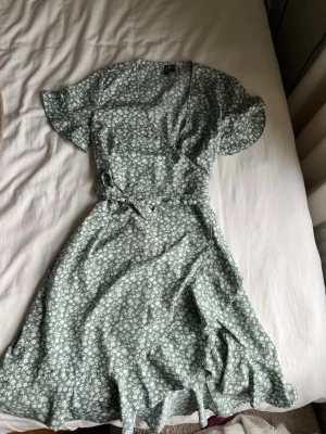 Grön blommig omlottklänning Vero Moda  - Söt grön omlottklänning från Vero Moda med vitt blommönster och knytband i midjan. Klänningen har korta ärmar och v-ringad hals. Perfekt för varma dagar och ger en fräsch sommarkänsla. Tunnt material!