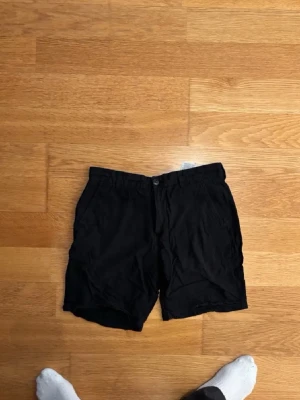 Svarta Jack and Jones shorts - Säljer ett par svarta shorts med normal passform och knappgylf. Shortsen har sidofickor fram och två bakfickor med knapp. Perfekta till sommaren och enkla att matcha med olika stilar. Materialet känns lätt och bekvämt.