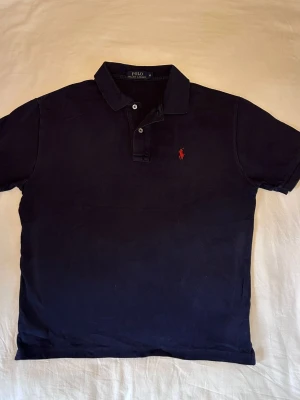 Ralph Lauren | Piké | M - Klassisk marinblå polotopp från Ralph Lauren med korta ärmar och krage. Toppen har knappar framtill och den ikoniska röda loggan broderad på bröstet. Tillverkad i bomull för en skön och stilren känsla. Mått: 63 Längd 52 Bredd