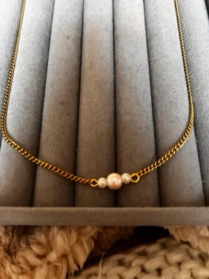 Guldhalsband med pärlor - Snyggt halsband med tunn guldfärgad kedja och tre små pärlor i mitten, varav en är ljusrosa och två vita. Perfekt för dig som gillar minimalistiska smycken med en touch av elegans. Passar till många olika stilar och är lätt att matcha med andra accessoarer.