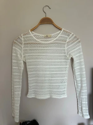 Vit spetsig långärmad topp Hollister - Säljer en vit långärmad topp från Hollister i spetsig, transparent design. Toppen har rund halsringning och ett fint mönster i spets som ger en luftig och trendig look. Perfekt att styla med linne under eller som lager på lager.