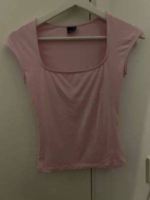 Gina Tricot - Söt ljusrosa topp med fyrkantig ringning och korta ärmar. Toppen har en slim passform och är tillverkad i ett mjukt, stretchigt material som känns skönt mot huden. Perfekt att styla med jeans eller kjol för en fräsch look.