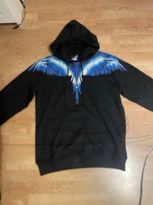 Marcelo burlon hoodie - Säljer en svart hoodie från Marcelo Burlon med coolt blått och vitt vingmotiv över axlarna och ryggen. Hoodien har huva med dragsko och är tillverkad i mjukt material. Perfekt för dig som vill sticka ut med en unik design.