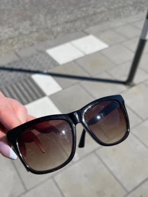 Svarta solglasögon från Gucci - Snygga svarta solglasögon från Gucci med fyrkantig form och mörktonade glas. Bågarna har diskret Gucci-logga och klassiska gröna och röda detaljer på sidan. Perfekta för att lyfta vilken outfit som helst. Fodral och äkthetskort medföljer!