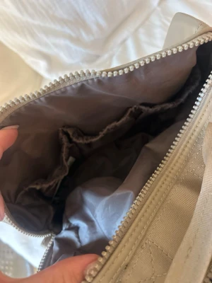 Necessär - Rymlig beige necessär med flera innerfack och dragkedja. Insidan är fodrad i ljusgrått tyg och har elastiska fickor för enkel organisering. Necessären har en praktisk bärrem upptill och är tillverkad i slitstarkt tygmaterial.