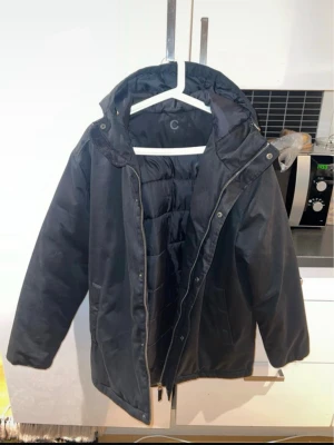 Svart pufferjacka med huva - Svart pufferjacka från C med huva och dragkedja framtill. Jackan har quiltad insida och en klassisk, rak passform som ger en clean look. Perfekt för kalla dagar när du vill hålla dig varm och stilsäker.