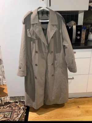 Beige trenchcoat från No 1 - Stilren beige trenchcoat från No 1 med klassisk dubbelknäppning och bred krage. Jackan har långa ärmar, rutigt innerfoder och bälte i midjan för en snygg siluett. Perfekt för dig som vill ha en tidlös look med cool attityd.