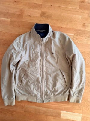 Gant New Hampshire Beige vindjacka - Stilren beige vindjacka från Gant med dragkedja framtill och två snedställda fickor med dragkedja. Jackan har ståkrage med mörkblå insida och diskret Gant-logga broderad på bröstet. Tillverkad i lätt material som passar perfekt för blåsiga dagar.