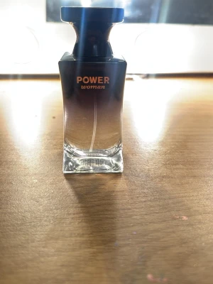Power Woman parfymflaska - Snygg parfymflaska från Power Woman med elegant svart och transparent design. Flaskan har en fyrkantig form med bred svart kork och text i guld. Perfekt för dig som gillar stilrena och moderna dofter.Nästan fylld ända upp.