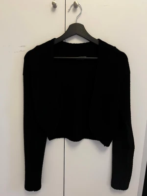 Svart kort kofta från Impulse - Snygg svart kort kofta från Impulse London. Koftan har öppen front, långa ärmar och ribbade detaljer vid hals och ärmslut. Perfekt att slänga över en topp för en enkel men stilren look.