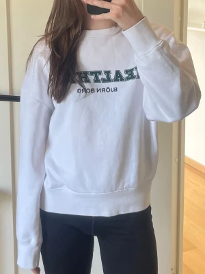 Vit sweatshirt från Björn Borg - Vit sweatshirt från Björn Borg med trycket 'HEALTHISM' i grönt och svart på bröstet. Tröjan har rund halsringning, ribbade muddar vid ärmslut och nederkant samt en avslappnad passform. Perfekt för en sportig och casual stil.