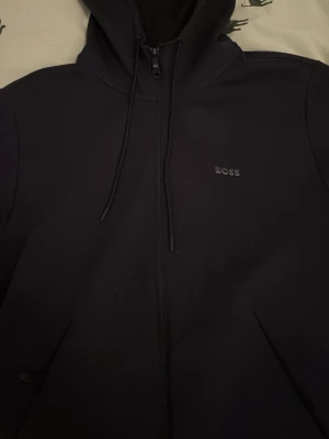 Navy hoodie från BOSS med dragkedja - Snygg navy hoodie från BOSS med dragkedja framtill och justerbar huva med snörning. Diskret BOSS-logga på bröstet. Perfekt för en clean och stilren look.