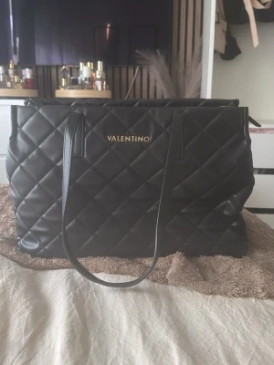 Svart quiltad handväska Valentino - Stilren svart handväska från Valentino med quiltat mönster och långa axelband. Väskan har en rektangulär form och är tillverkad i vadderat konstläder. Guldigt Valentino-tryck framtill ger en lyxig touch. Perfekt för dig som gillar klassisk och trendig design.