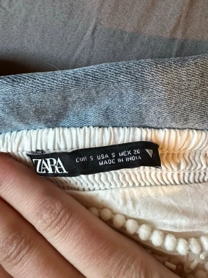 Vit kort klänning från Zara - Säljer en söt vit kort klänning från Zara med volangkant och små dekorativa bollar längs nederdelen. Klänningen har en avslappnad passform och resår i midjan. Perfekt för sommardagar eller när du vill känna dig extra lätt och fri.