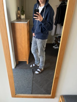 Mörkblå zip-hoodie  - Snygg mörkblå hoodie med dragkedja och vita snören i huvan. Klassisk modell med fickor framtill och ribbade muddar vid ärmslut och nederkant. Perfekt för en avslappnad stil och enkel att matcha med jeans eller joggers.