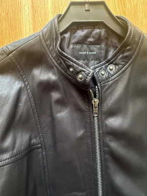 Svart skinnjacka från Samsøe Samsøe - Skinnjacka by Samsøe Samsøe från Danmark  Väldigt bra skick som ny  Size 2XL men kan vara som XL om du har breda axlar .  Äkta läder . SIZE: Bredden under armhålorna : 56cm Armlängden från armhålan : 68cm  Längd : 74cm