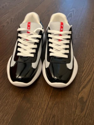 Svarta och vita sneakers från Prada - Snygga sneakers från Prada i svart och vitt med glansiga svarta detaljer, vit mesh och röda loggor på plös och häl. Skorna har vita snören och en chunky vit sula. Perfekta för dig som vill sticka ut med en lyxig och modern look.