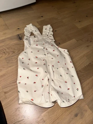 Vit blommig jumpsuit från H&M - Vit blommig jumpsuit från H&M Supersöt vit jumpsuit med små röda och gröna blommor från H&M. Axelbanden har volangdetaljer och knäpps med tryckknappar. Jumpsuiten har en ficka framtill och är tillverkad i mjukt bomullstyg. Perfekt för en lekfull och trendig look.