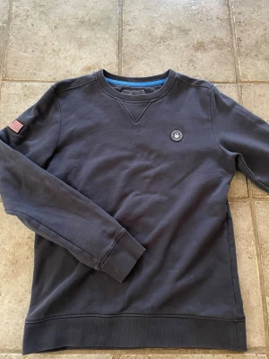 Mörkblå sweatshirt från Sail Racing - Mörkblå sweatshirt från Sail Racing med rund halsringning, ribbade muddar och en liten amerikansk flagga på ärmen. Tröjan har en diskret logotyp på bröstet och är tillverkad i mjukt bomullsmaterial.
