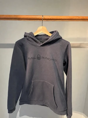 Sail Racing hoodie  - Hoodie från Sail Racing med stor känguruficka framtill och tryckt logga på bröstet. Klassisk huva och mjukt material som känns skönt mot huden. Perfekt för en avslappnad och sportig stil. Diskret logga även på huvan baktill.