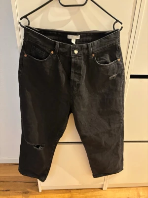 Svarta slitna jeans från H&M - Säljer ett par svarta jeans från H&M med avslappnad passform och slitningar på ena benet. Jeansen har klassisk femficksdesign, hög midja och raka ben. Perfekta för en cool och trendig look.