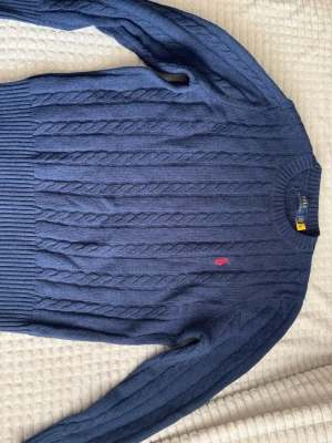 Mörkblå kabelstickad tröja Polo Ralph Lauren Strolek L men sitter som M - Mörkblå kabelstickad tröja från Polo Ralph Lauren med klassisk rund halsringning och röd broderad logga på bröstet. Tröjan har långa ärmar och ribbstickade muddar. Perfekt för dig som gillar stilren och tidlös design. säljer eftersom den ej passar mer storlek L men sitter som M