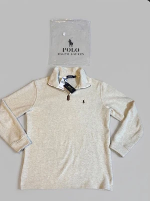  Polo Ralph Lauren 1/4 zip L - Helt ny Stilren beige långärmad tröja från Polo Ralph Lauren med halvzip och hög krage. Tröjan har det klassiska broderade logotypen på bröstet och är tillverkad i mjukt material. Perfekt för en avslappnad men snygg look.