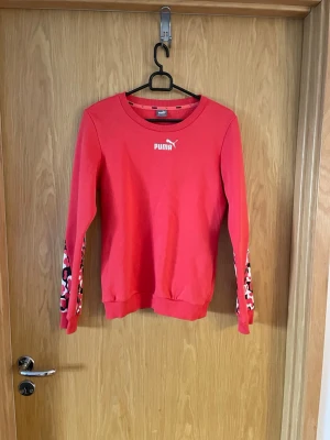 Röd tjock  tröja från Puma - Röd tjock tröja från Puma med rund halsringning och långa ärmar. Ärmarna har coolt mönster i svart, vitt och rosa längs sidorna. Tröjan har ribbade muddar vid ärmslut och nederkant samt vit Puma-logga på bröstet.