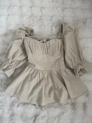 Beige topp  med puffärmar - Supersöt beige topp med puffiga långärmar och markerad midja. Toppen har fina veckdetaljer över bysten, knytband i ryggen och en lätt utställd kjol. Tillverkad i ett linneliknande material som känns luftigt och perfekt för varma dagar.