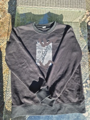 Svart sweatshirt med grafiskt tryck - Cool svart sweatshirt med ett grafiskt tryck av ett ansikte i vitt och svart på bröstet. Tröjan har rund halsringning och ribbade muddar vid ärmslut och nederkant. Perfekt för en avslappnad streetwear-look. Riktiga bilder går att lösa 