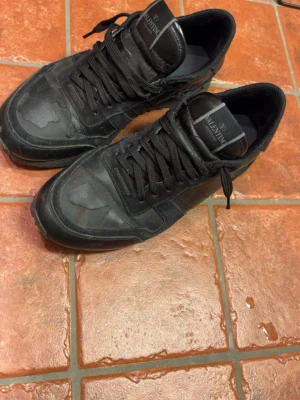 Svarta Valentino sneakers i läder - Snygga svarta sneakers från Valentino med coola detaljer och robust sula. Skorna är gjorda i en mix av läder och mocka, med svarta skosnören och diskret logga på plösen. Perfekta för dig som gillar stilrena och exklusiva sneakers.
