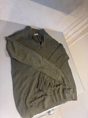 Olivgrön halvzip tröja mango  - Olivgrön långärmad tröja med halv dragkedja vid halsen. Tröjan har ribbade muddar vid ärmslut och nederkant, vilket ger en snygg och avslappnad look. Perfekt för lager-på-lager-stil under kyliga dagar.