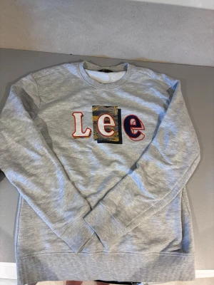 Grå sweatshirt från Lee - Cool grå sweatshirt från Lee med broderad logga i rött, vitt och blått på bröstet. Tröjan har rund halsringning, ribbade muddar och är tillverkad i mjukt material som passar perfekt för en avslappnad look.